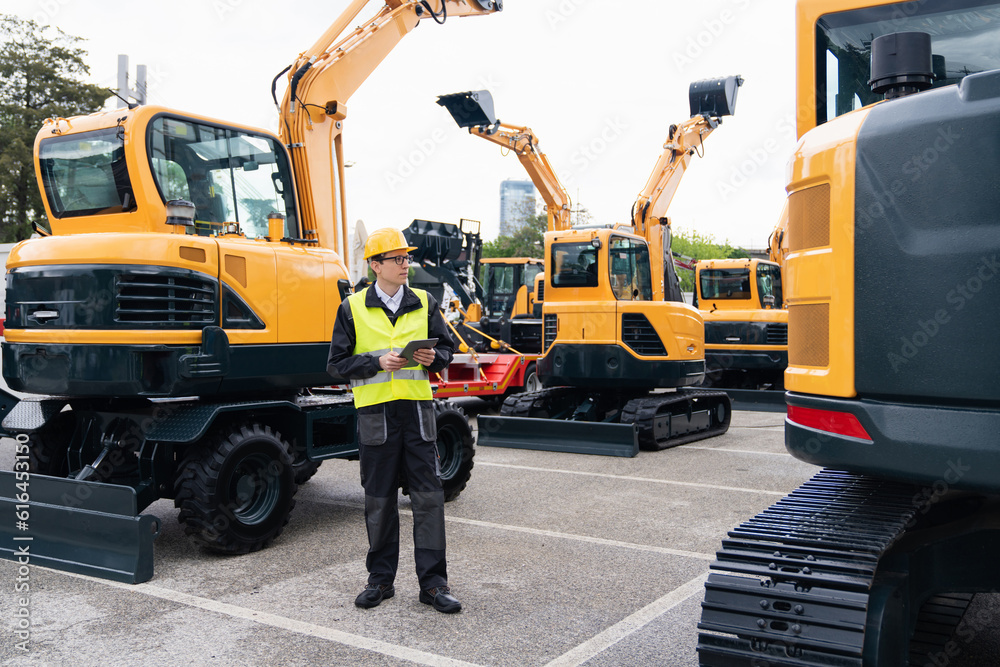 Engineer in a helmet with a digital tablet stands next to construction excavators.