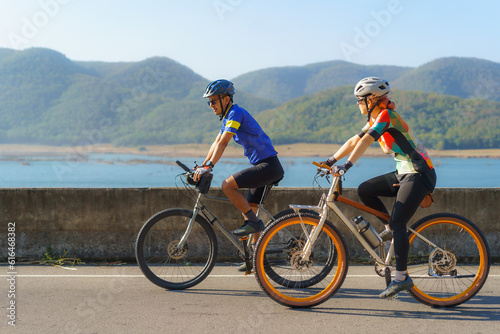 Asian cyclist couple riding together for exercise around the lake in the morning with beautiful mountain view in the background...