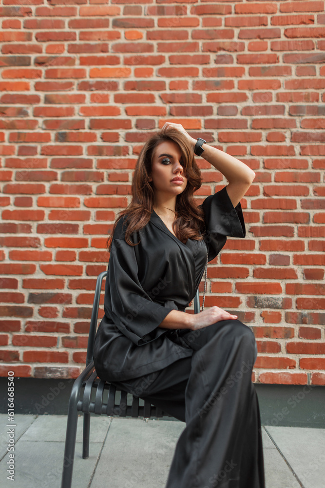 Beautiful elegant young stylish woman in a fashionable black suit with pants and a shirt sits near a red brick wall on the street. Pretty lady