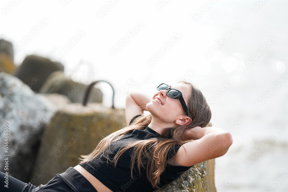 A beautiful young girl in sunglasses is lying on a rock on the seashore