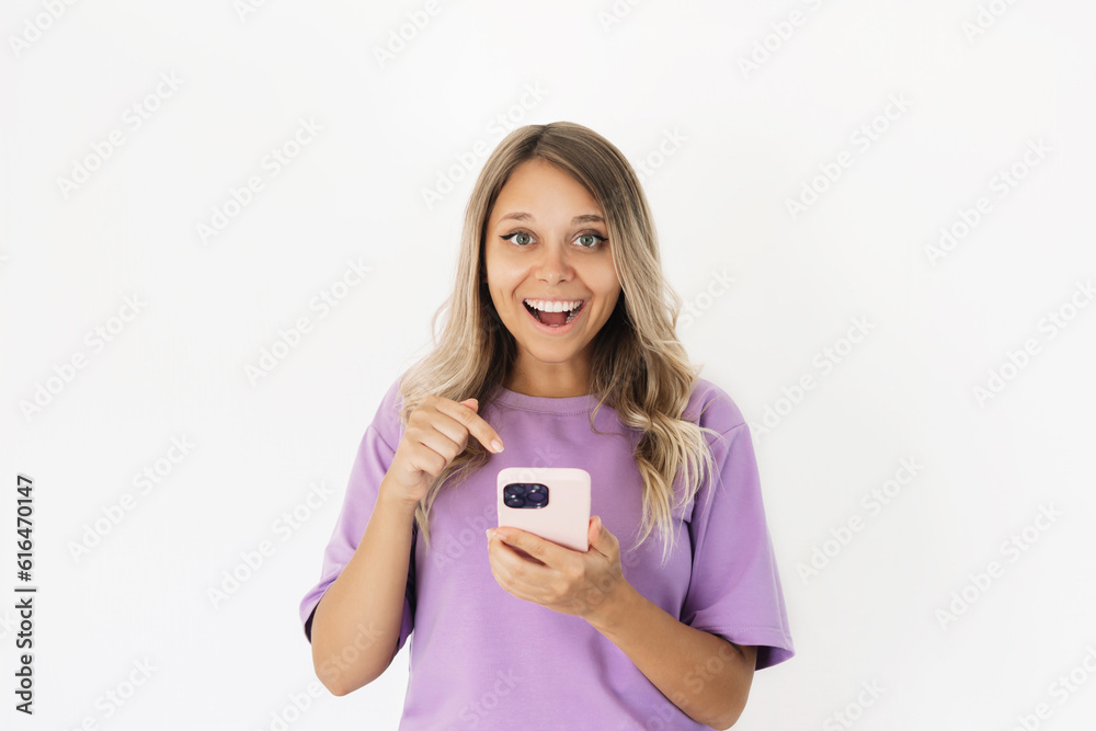 A young attractive caucasian impressed excited smiling blonde woman in a violet t-shirt holds mobile phone in her hand and points at the screen with her finger isolated on a white background A young attractive caucasian impressed excited smiling blonde woman in a violet t-shirt holds mobile phone in her hand and points at the screen with her finger isolated on a white background