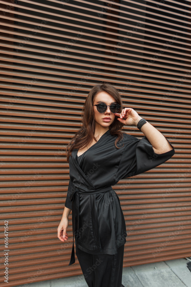 Glamorous beautiful fashion chic young woman model with brunette hair in a fashionable elegant black suit against the background of a metal rusty wall in the city Glamorous beautiful fashion chic young woman model with brunette hair in a fashionable elegant black suit against the background of a metal rusty wall in the city