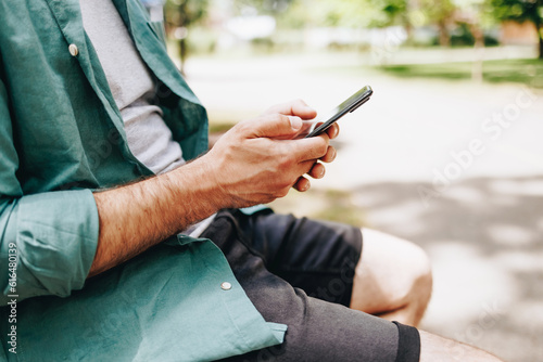 Happy young handsome man sitting on the bench outdoors and using smartphone