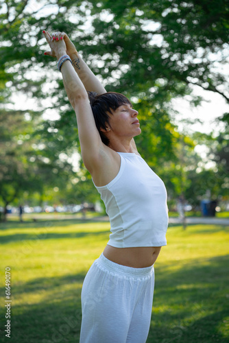 Full length of smiling young sporty girl in white sportswear standing outside against background of green lush trees in city park, female during yoga class in nature