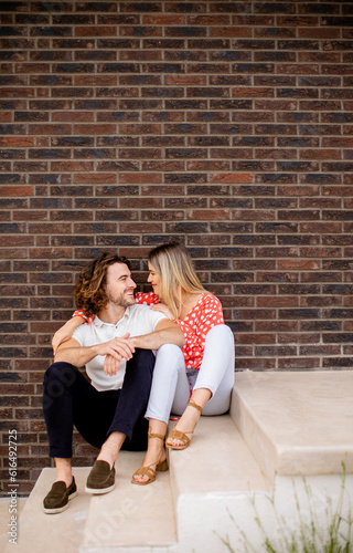 Smiling young couple in love sitting in front of house brick wall