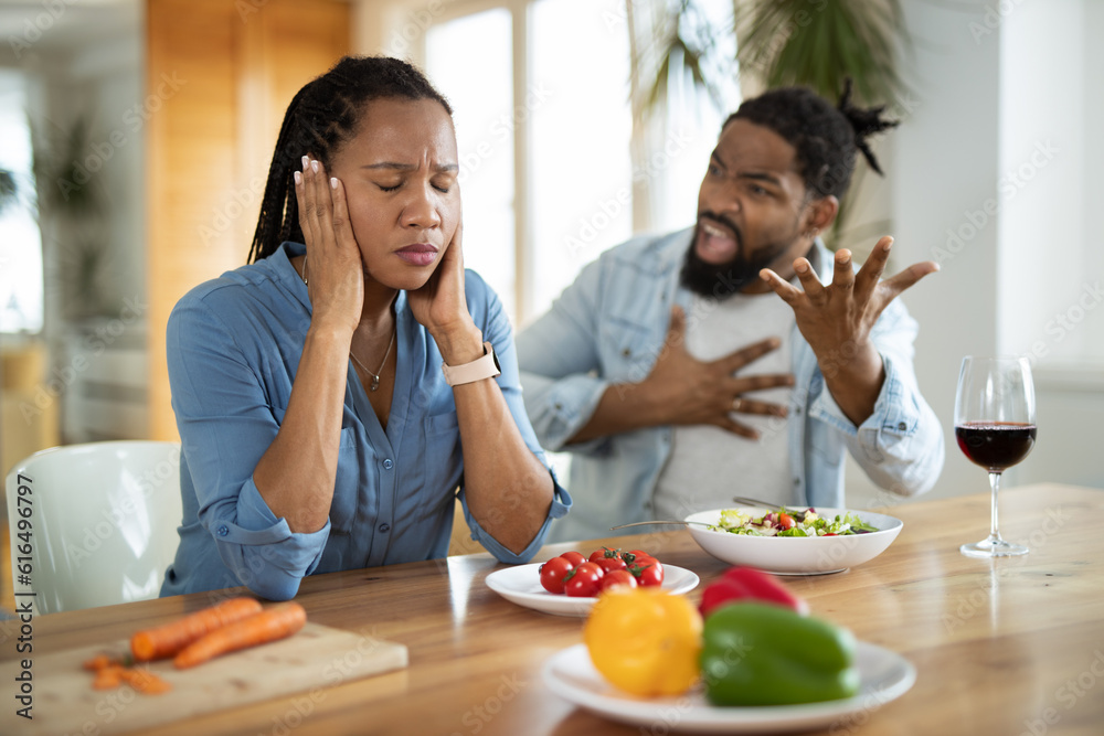 Angry black man shouting at his wife during their breakfast at dining table while she is covering her ears