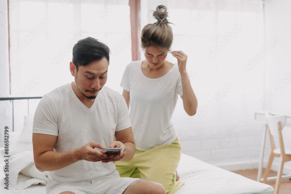 Fototapeta premium Funny asian couple. Wife peeking husband who chatting on the smartphone.