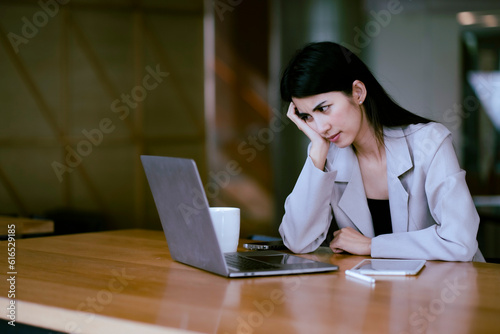 Young Asian businesswoman stress with her work, looking at notebook with hand under chin and folding eye bow, electronic device on tavle with copy space. photo