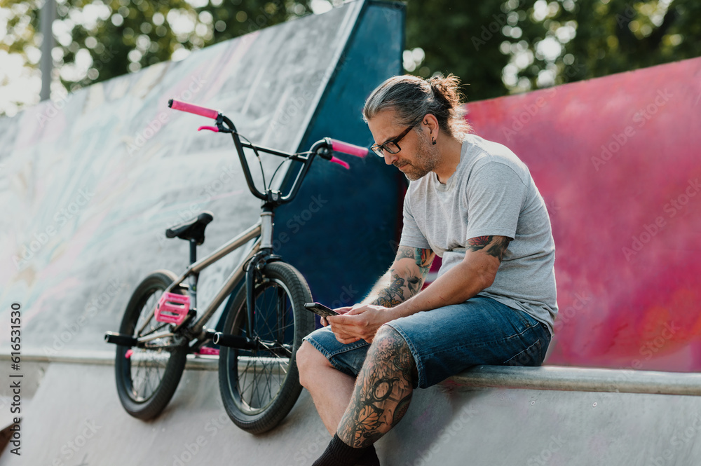 Obraz premium A serious urban tattooed middle-aged man is sitting in a skate park next to his bmx bike and scrolling on his phone.