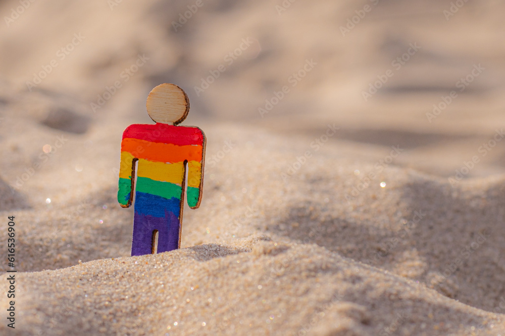 Naklejka premium LGBT wooden people on the beach on the sand. Couple standing on beach with rainbow flag, equal rights, symbol of LGBT community. LGBT flag on a sandy beach. Summer travel concept. A symbol of same-sex