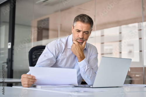 Doubtful serious busy mid aged mature professional business man ceo manager executive holding corporate financial documents reading paper letter sitting at desk in office feeling doubt uncertainty.