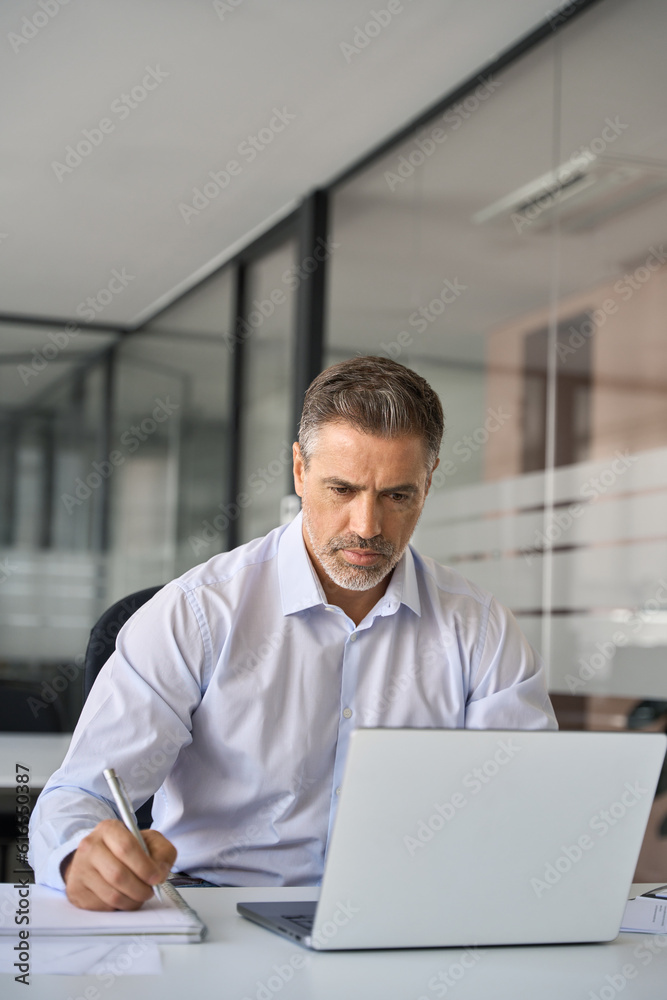 Busy serious business man executive of mid age sitting at desk working on laptop writing notes. Professional businessman manager using computer elearning watching webinar in modern office. Vertical