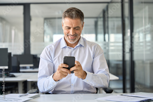 Happy older mid aged business man ceo executive, mature businessman manager sitting in modern office holding smartphone using mobile cell phone managing digital tech transactions on cellphone at work.