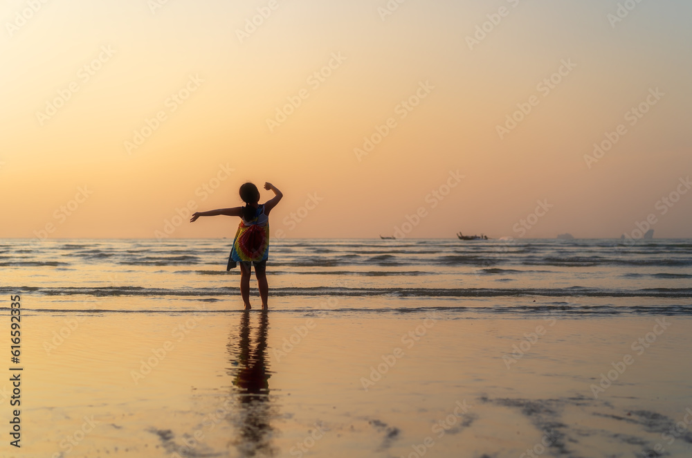 back asian child or kid girl standing on summer sand beach and sea to happy raising hands with golden warm sunlight on sunset twilight or morning sunrise for family vacation travel on holiday relax
