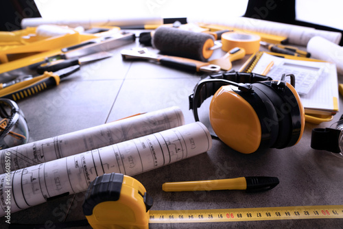 Contractor and construction concept. Tool kit of the contractor: yellow hardhat, hearing protection, libella, hand saw, and notebook on the gray tiles background.