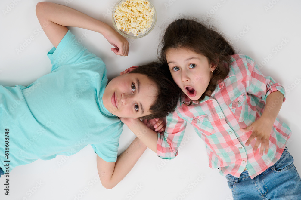 Happy children with popcorn bowl smile at camera, express surprise and amazement, lying on their backs on isolated white studio background. Kids. Childhood. Entertainment. Lifestyle. Leisure activity