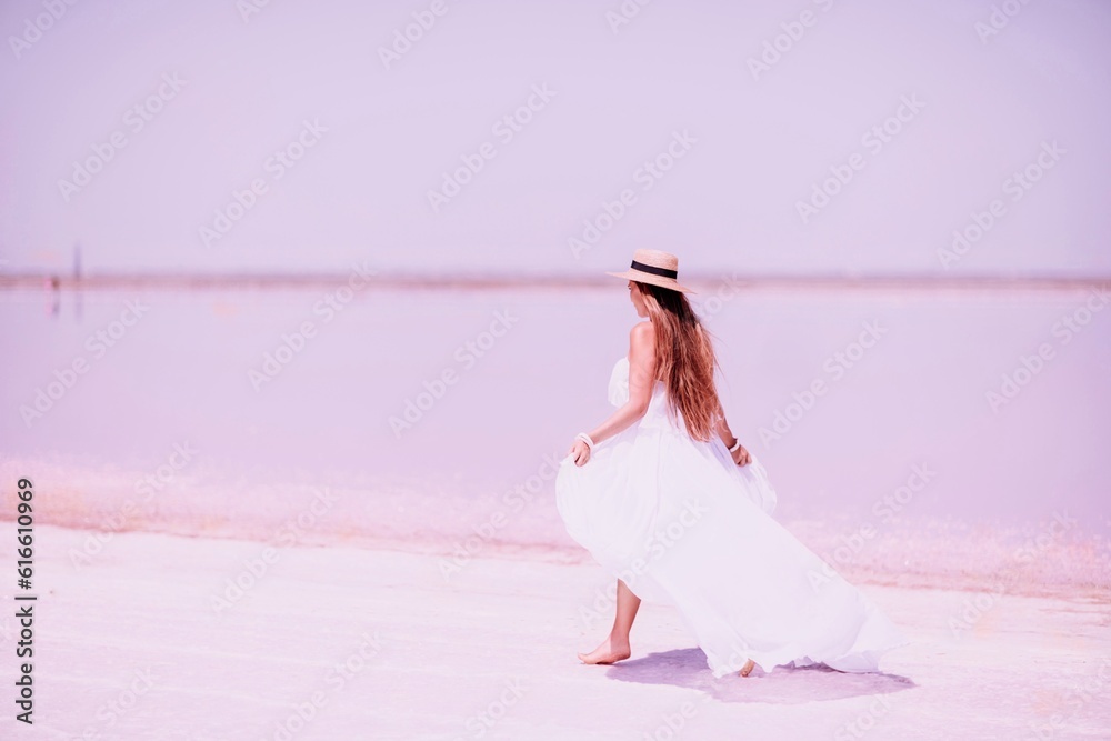 Naklejka premium Woman in pink salt lake. She in a white dress and hat enjoys the scenic view of a pink salt lake as she walks along the white, salty shore, creating a lasting memory.