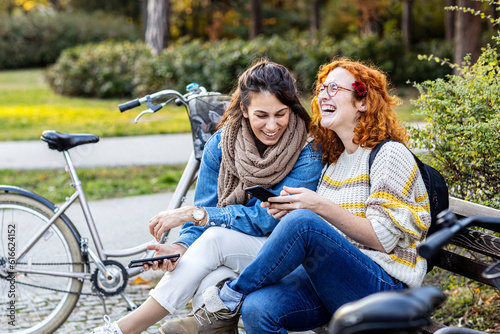 Two girl friends sitting on bench, looking at mobile phone and smile in public par with bycicle on background