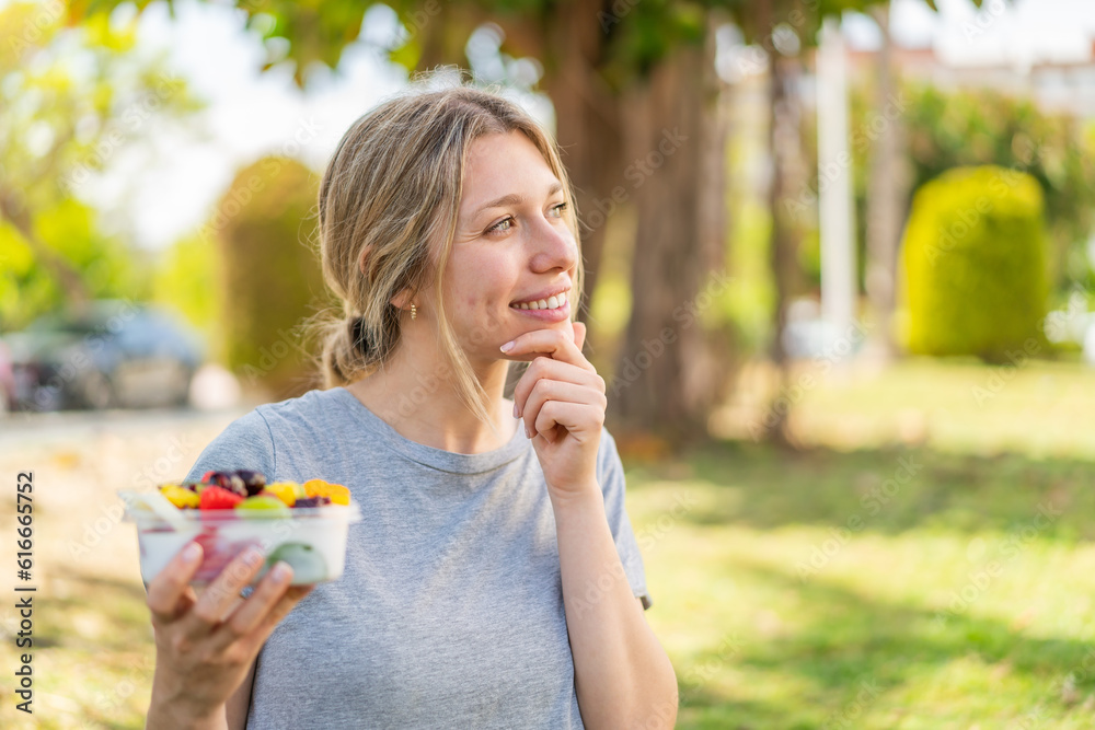 Fototapeta premium Young blonde woman holding a bowl of fruit at outdoors thinking an idea and looking side