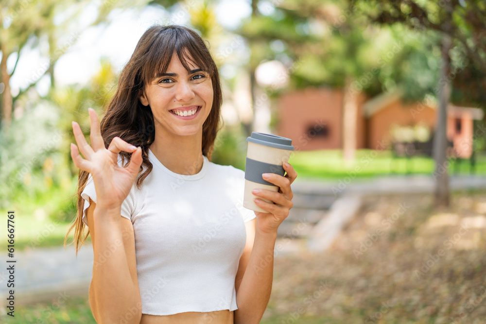 Young woman holding a take away coffee at outdoors showing ok sign with fingers