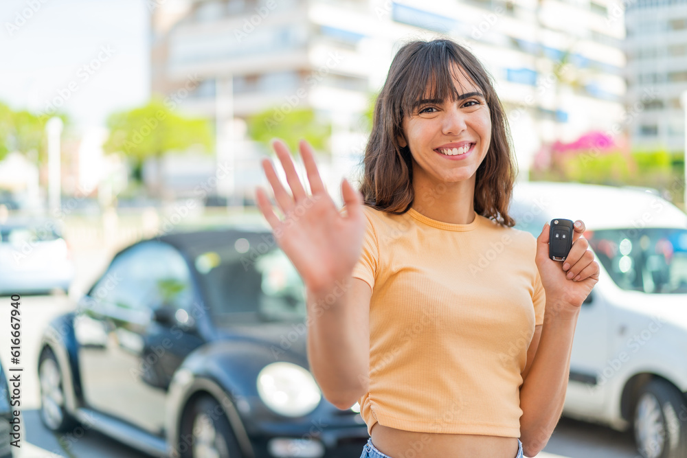 Young woman holding car keys at outdoors saluting with hand with happy expression