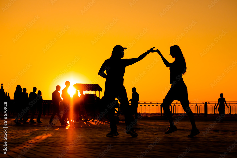 Man and woman couple silhouettes dancing against warm sunset orange sky on quay at evening - sun lens flare. Group dance, romantic, love, summer outdoor activity and vacation concept Man and woman couple silhouettes dancing against warm sunset orange sky on quay at evening - sun lens flare. Group dance, romantic, love, summer outdoor activity and vacation concept