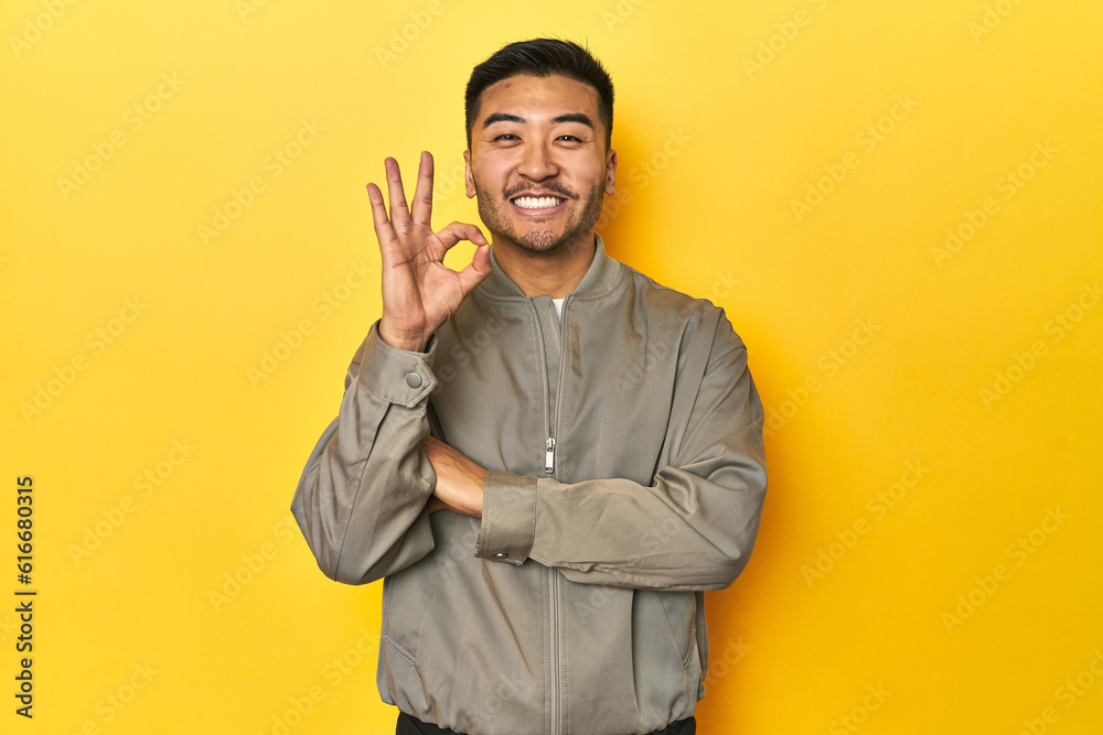 Stylish Asian man in gray jacket on yellow studio winks an eye and holds an okay gesture with hand.