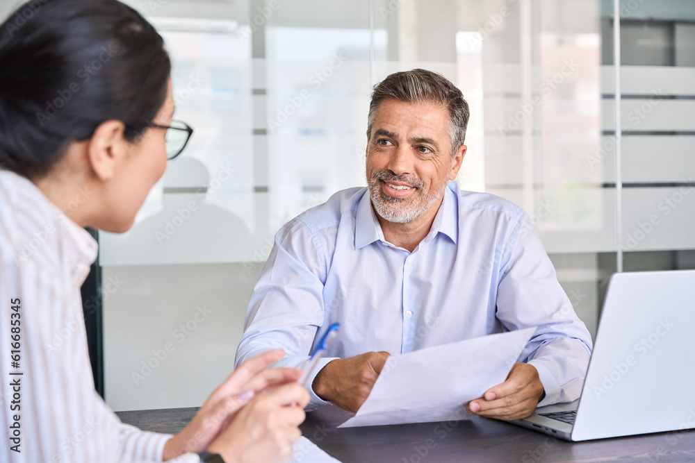 Smiling professional Latin employer hr manager holding cv during interview hiring for job female Asian female recruit, advisor consulting client sitting in office at meeting. Human resources. Smiling professional Latin employer hr manager holding cv during interview hiring for job female Asian female recruit, advisor consulting client sitting in office at meeting. Human resources.