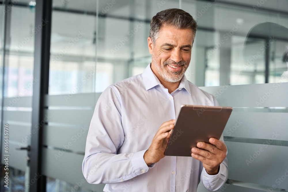 Young smiling busy mature male manager using tablet standing in office. Older mid aged professional business man company executive holding tab digital computer managing financial banking data. Young smiling busy mature male manager using tablet standing in office. Older mid aged professional business man company executive holding tab digital computer managing financial banking data.