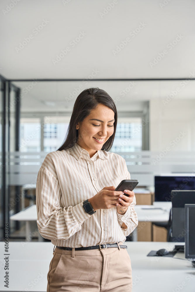 Young happy smiling professional Asian business woman executive manager employee holding cellphone using mobile phone standing in office hall working on smartphone, vertical.