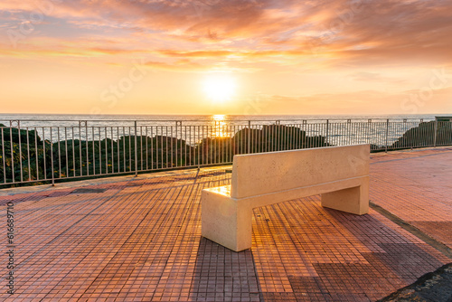 beautiful white stone bench on seafront embarkment during beautiful sunrise or sunset with sidewalk with pavement, sea surf and nice cloudy sunset sky © Yaroslav