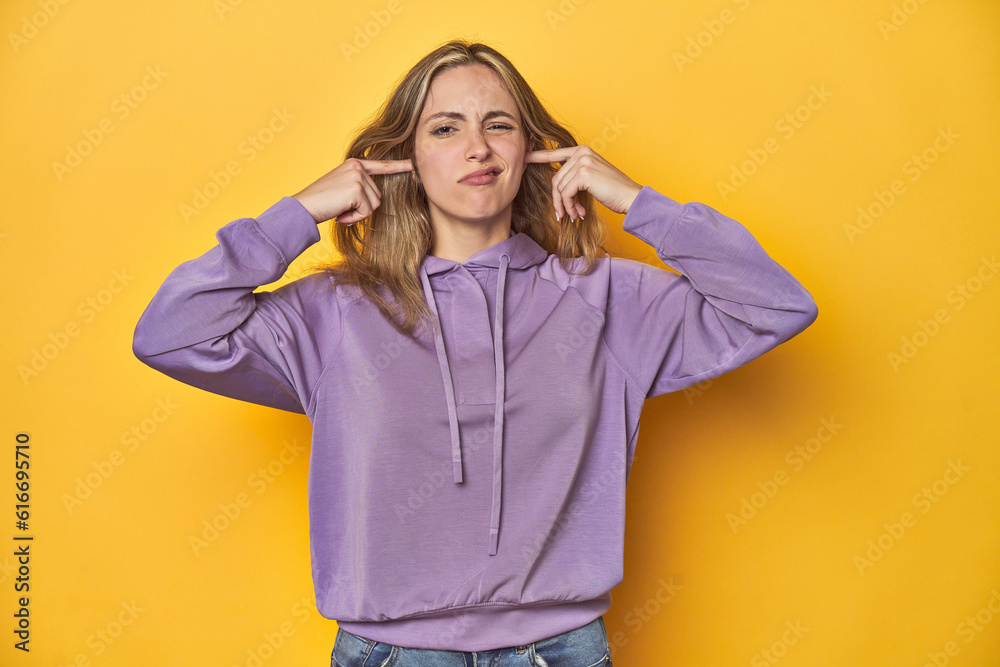 Young blonde Caucasian woman in a violet sweatshirt on a yellow background, covering ears with fingers, stressed and desperate by a loudly ambient. Young blonde Caucasian woman in a violet sweatshirt on a yellow background, covering ears with fingers, stressed and desperate by a loudly ambient.