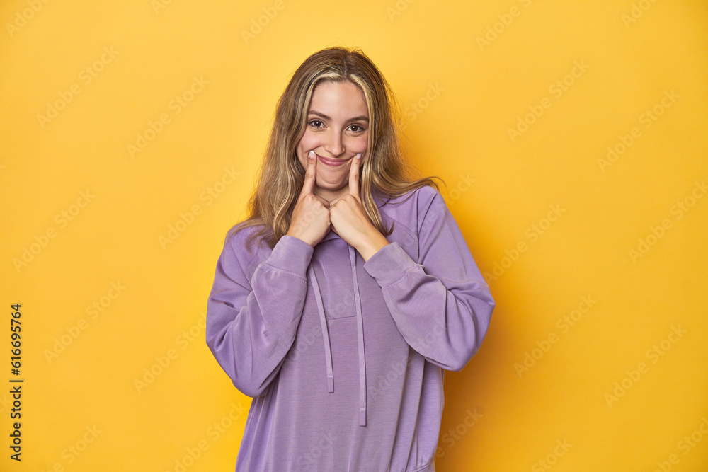 Young blonde Caucasian woman in a violet sweatshirt on a yellow background, doubting between two options. Young blonde Caucasian woman in a violet sweatshirt on a yellow background, doubting between two options.