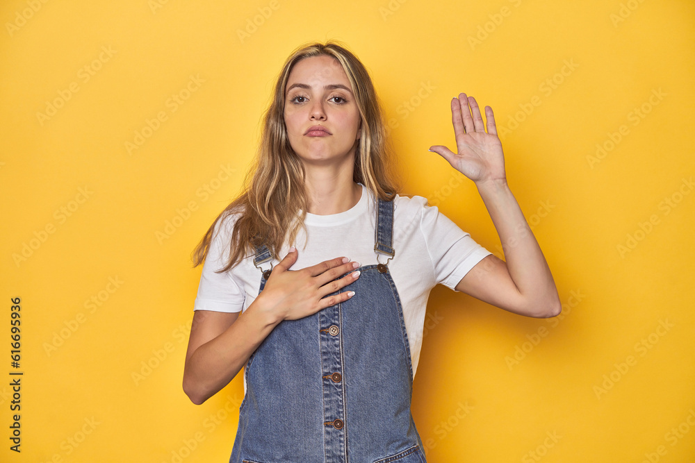 Young blonde Caucasian woman in denim overalls posing on a yellow background, taking an oath, putting hand on chest.