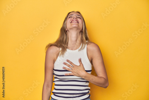 Young blonde Caucasian woman in a white tank top on a yellow studio background, laughs out loudly keeping hand on chest.