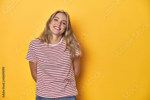Young blonde Caucasian woman in a red striped t-shirt on a yellow background, happy, smiling and cheerful.