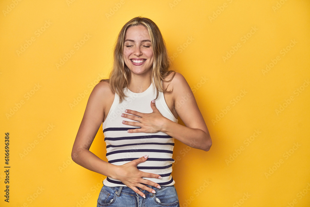 Young blonde Caucasian woman in a white tank top on a yellow studio background, laughs happily and has fun keeping hands on stomach. Young blonde Caucasian woman in a white tank top on a yellow studio background, laughs happily and has fun keeping hands on stomach.