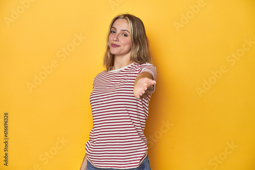 Young blonde Caucasian woman in a red striped t-shirt on a yellow background, stretching hand at camera in greeting gesture.