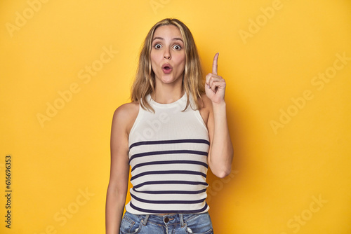 Young blonde Caucasian woman in a white tank top on a yellow studio background, having an idea, inspiration concept.