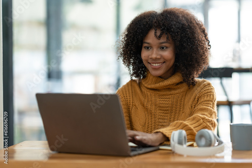 Young African businesswoman wearing headphones while looking at online shopping data Taking note of customer orders on a laptop while holding a mug of warm coffee to relax.