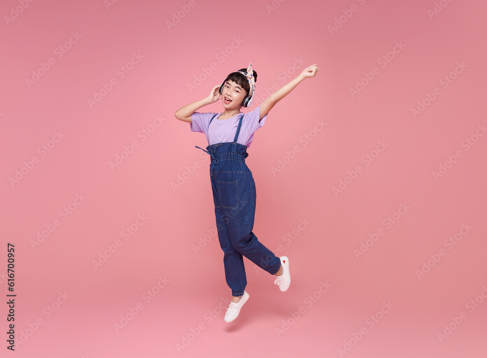 Full length happy asian teen children girl wearing headphones listening to music and jumping on pink studio background. People lifestyle relax