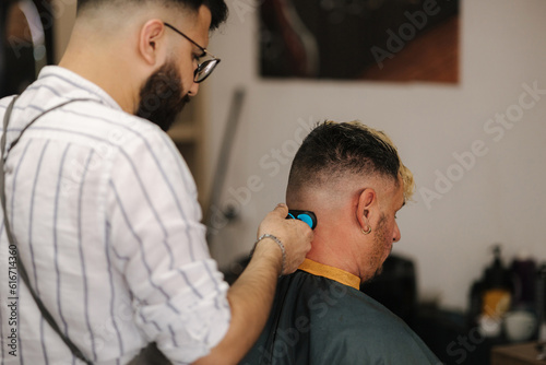 Bearded barber cutting hair of male customer. Hairstylist serving client at barber shop.