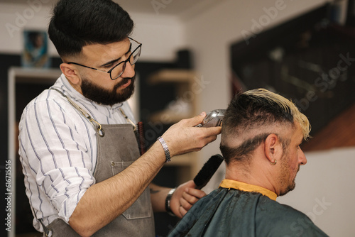 Bearded barber cutting hair of male customer. Hairstylist serving client at barber shop.