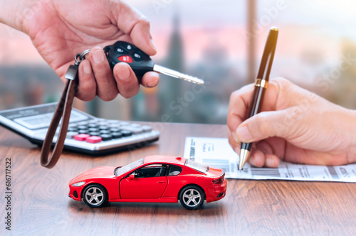 Car loan or buying a new car concept. Red car on table and salesman giving a car key to buyer  which is signing on paper in background. © Boxyray