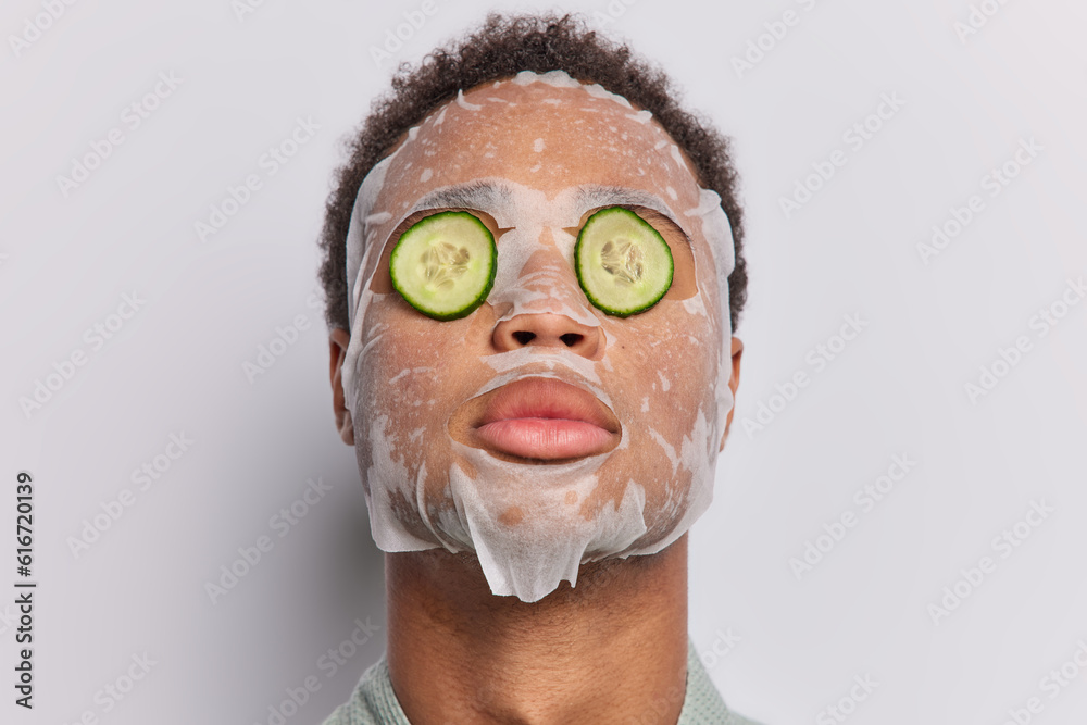Fototapeta premium Photo of dark haired man applies sheet facial mask covers eyes with cucumber slices undergoes daily procedures for rejuvenation isolated over white background. People wellness and skin care concept