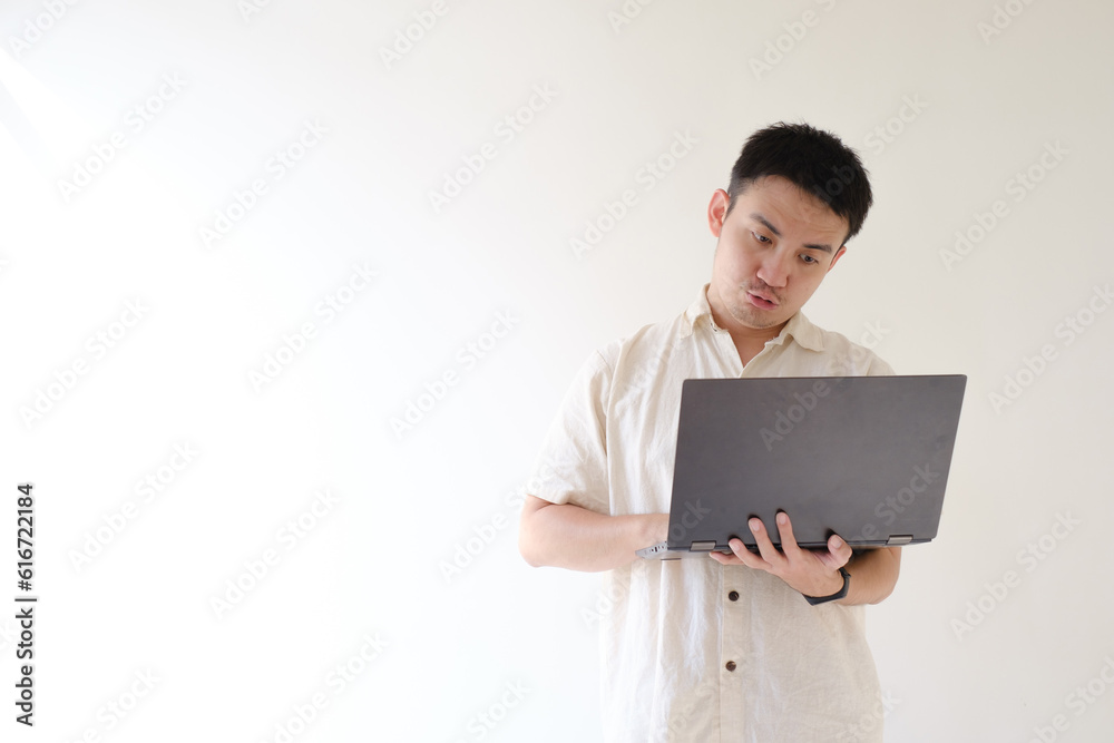 A young Asian man wearing a beige shirt and a smartwatch on his left wrist is using a black laptop withstanding and funny facial expression. Isolated white background. Suitable for advertisement.