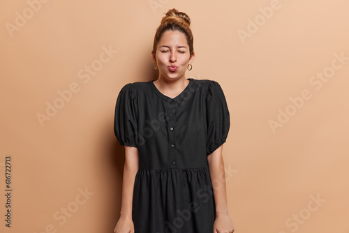 Pretty young woman with folded lips and expression of anticipation exudes flirty mood creating air of intrigue awaits kiss keeps eyes closed wears black dress isolated over brown background. © wayhome.studio 