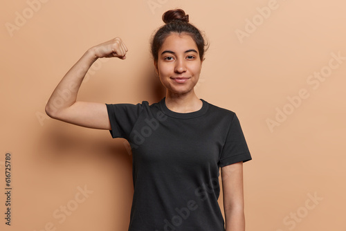 Self confident Iranian girl flexing her biceps and maintaining eye contact with camera demonstrates strength being strong after regular workout wears casual black t shirt isolated on brown background
