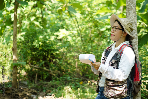 Asian woman botanist is at forest, hold paper notepad to survey  botancal plants in forest. Concept,  field research outdoor.                                  Nature surveying. Ecology and environment photo