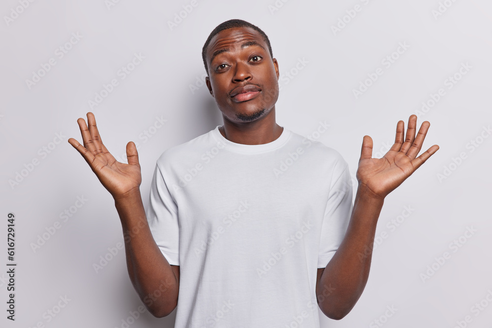 Fototapeta premium Indoor shot of hesitant dark skinned handsome man shrugs shoulders keeps palms raised up being doubtful dressed in casual t shirt isolated over white background. I dont know and cannot give you answer